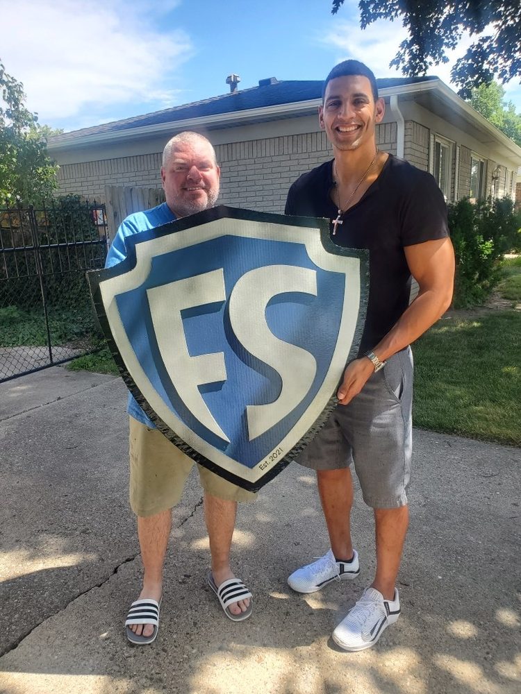 Custom FS shield logo cutout in blue, silver and black, Est. 2021, held by two men outside