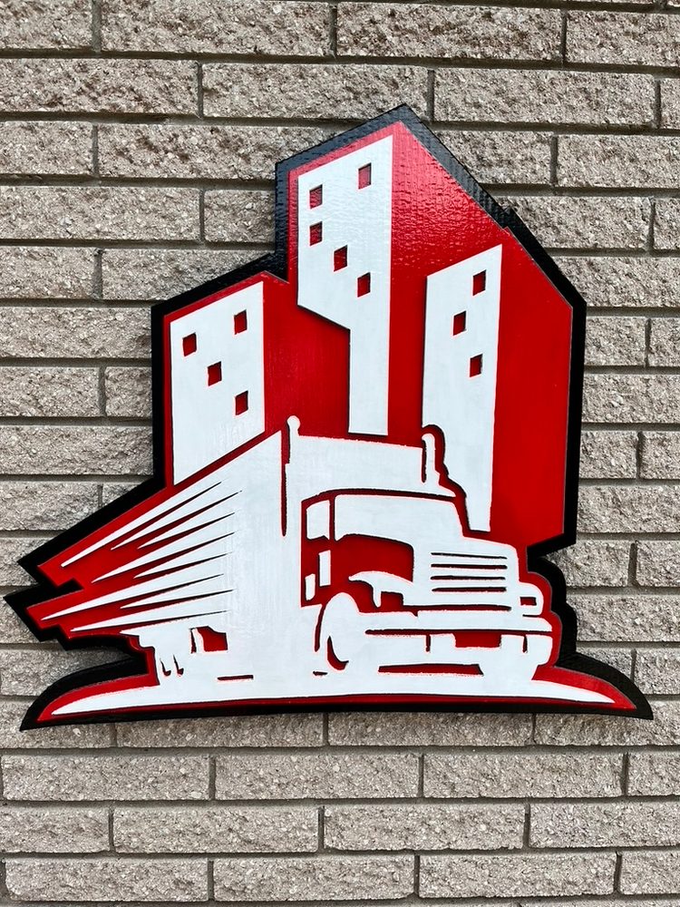 Custom red, white and black trucking company logo cutout of a semi truck in front of city buildings, mounted on a brick wall