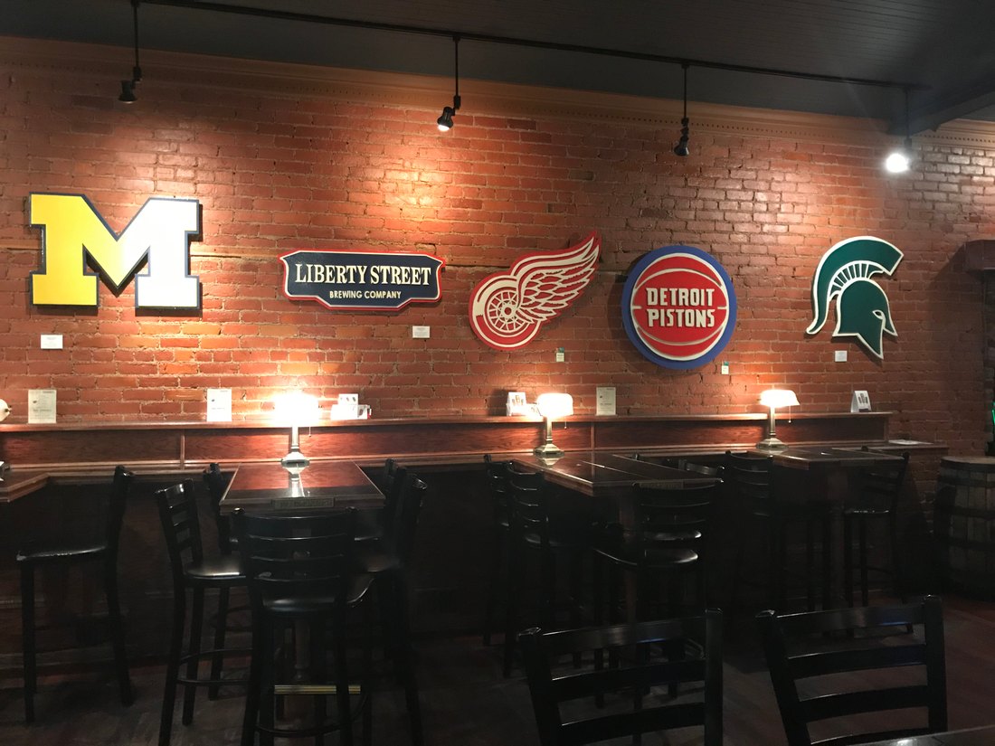 Custom Michigan block M, Liberty Street Brewing Company sign, Detroit Red Wings wing, Detroit Pistons logo and Michigan State Spartan helmet in a row on a brick wall at Liberty Street Brewing
