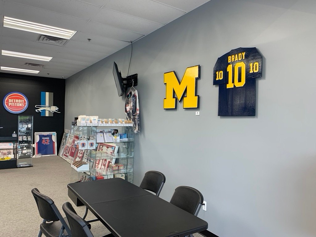 Custom Michigan block M and Brady number 10 tribute jersey cutouts on a shop wall, with the Detroit Pistons logo and Detroit Lions cutout on the far wall
