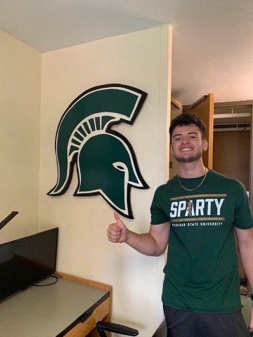A Michigan State student in a Sparty shirt giving a thumbs up next to a custom Spartan helmet cutout on his dorm room wall