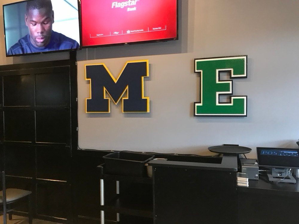 Custom navy Michigan block M with maize trim beside a green Eastern Michigan block E on a gray bar wall