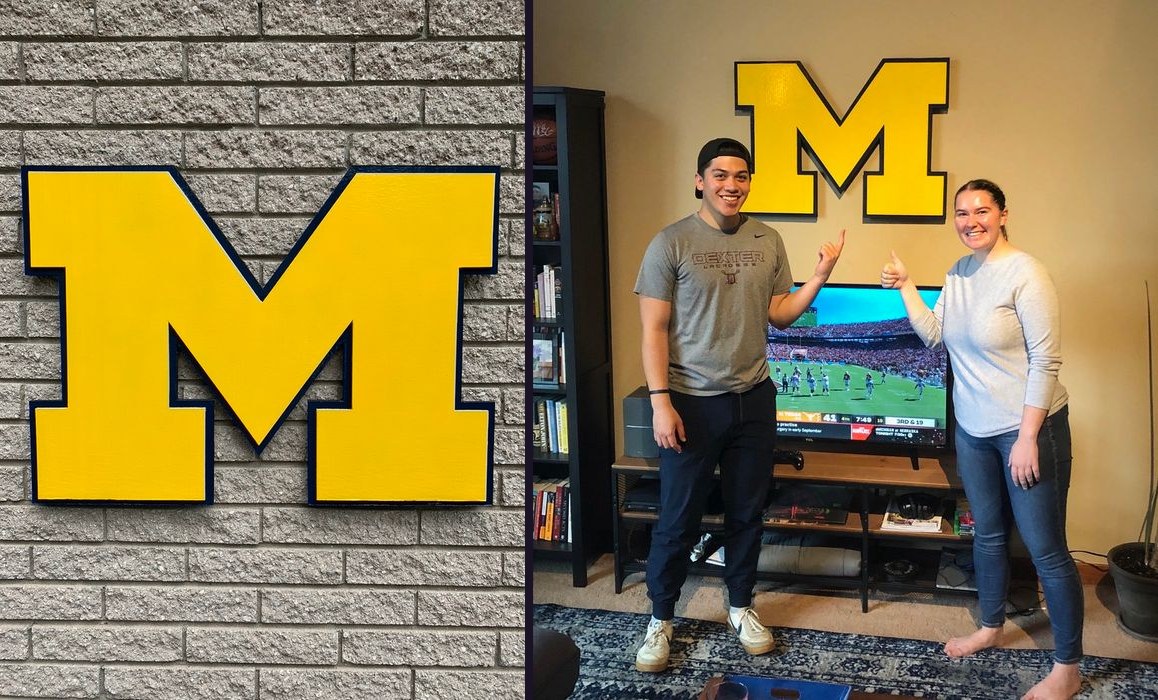 Custom maize Michigan block M cutout with navy trim on a brick wall, and a couple pointing at the maize M above their TV during a Michigan game