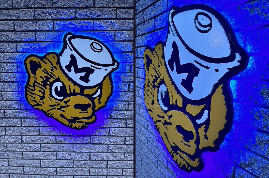 Custom vintage Michigan Wolverine cutout glowing with blue LED backlighting on a brick wall, front view and angled side view