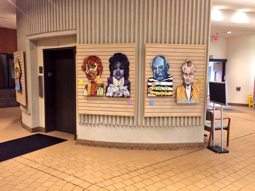 Four cardboard portraits hung on wood slat panels in the Livonia City Hall lobby