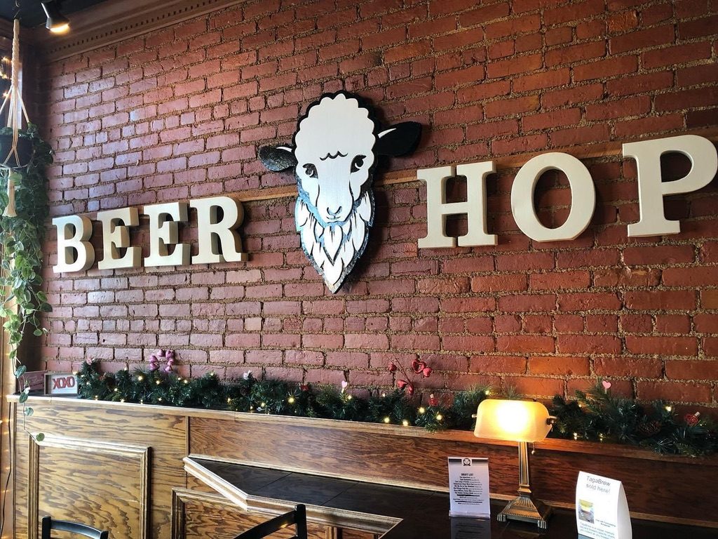 The hand-cut Bearded Lamb sheep logo on the brick wall between BEER and HOP lettering, with a lamp and garland below