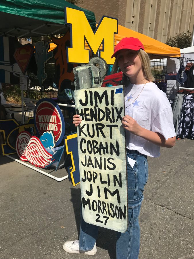 A customer holding a tall painted sign listing Jimi Hendrix, Kurt Cobain, Janis Joplin and Jim Morrison, 27, in front of the booth