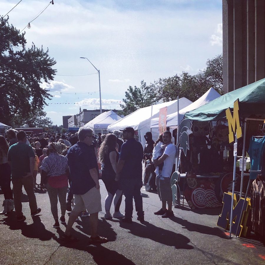 Festival-goers walking past tents and a booth with a Michigan M in the sunlight at Dally in the Alley