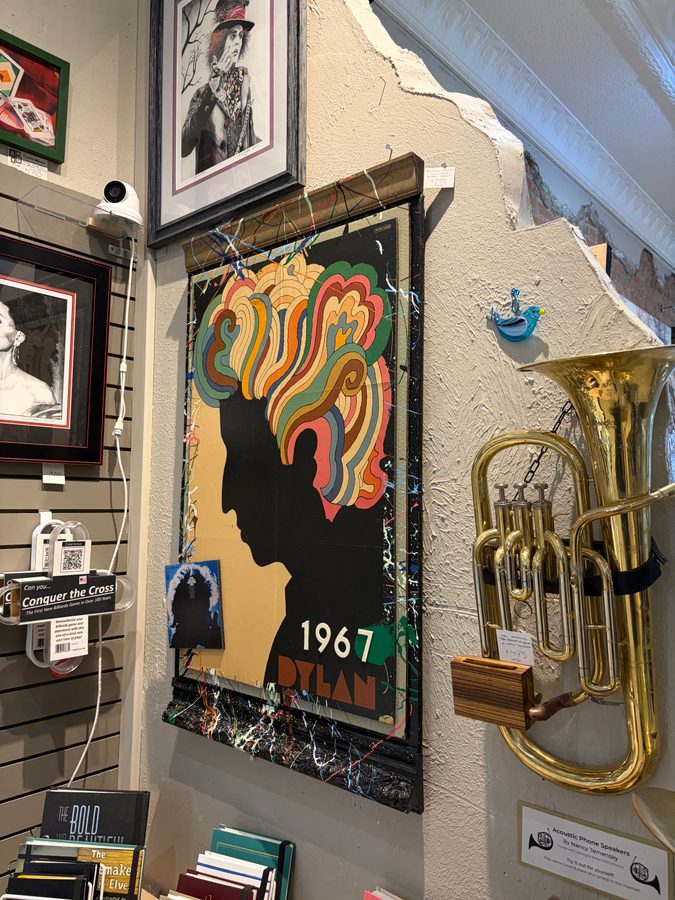 A framed Dylan 1967 piece with a silhouette and colorful hair on the wall at Frame Works next to a brass tuba and black and white portraits