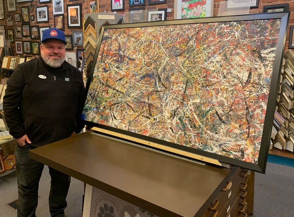 Jeremy Rheault standing beside the framed splatter painting Journey in the Frame Works shop, with frame samples on the wall behind