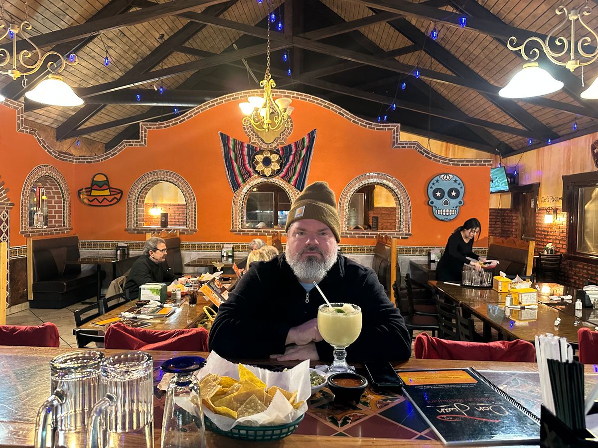 Jeremy Rheault seated at the bar at Los Amigos with chips and a margarita, the orange wall of pieces behind him