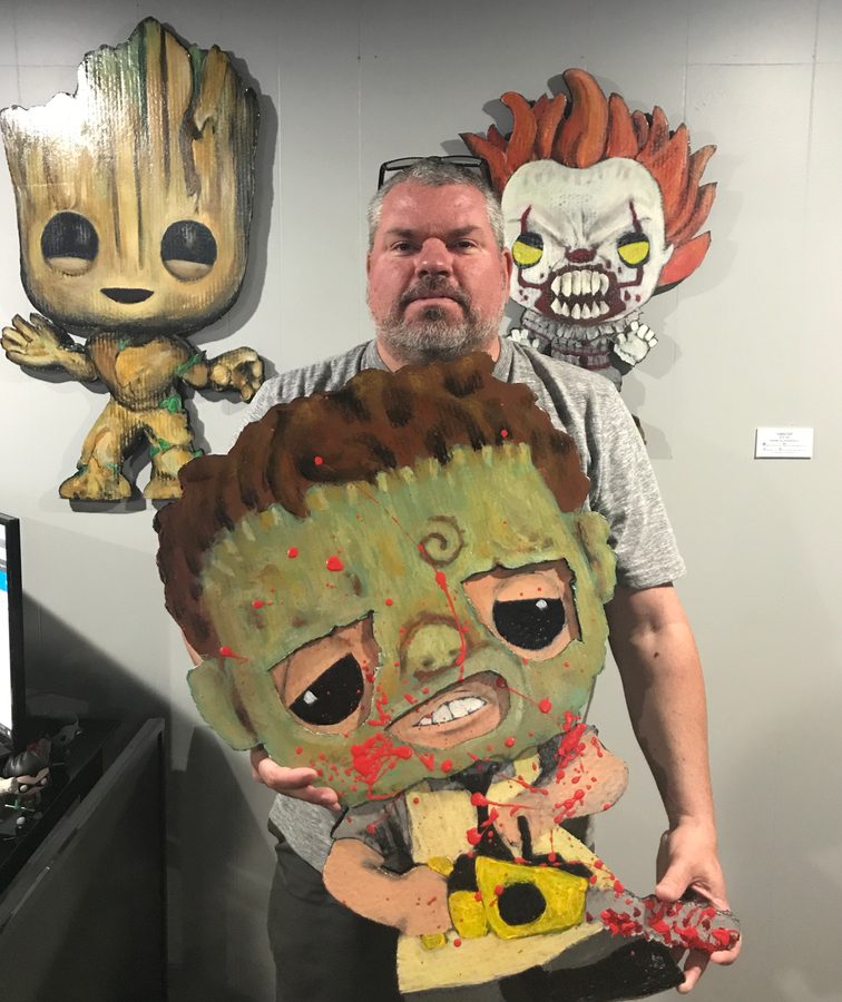 Jeremy Rheault holding a hand-painted Leatherface Funko-style cutout in front of Pennywise and Groot on a gallery wall