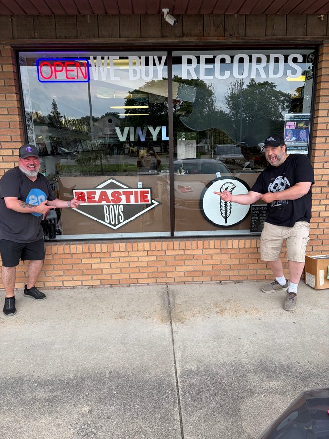 Two men posing at the front window of Vinyl People Records with the Beastie Boys and feather logo pieces