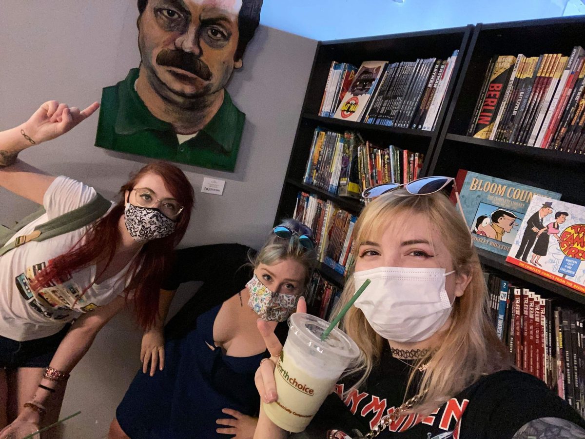 Three alumni in masks taking a selfie under my hand-painted Ron Swanson portrait on the wall at State of Comics